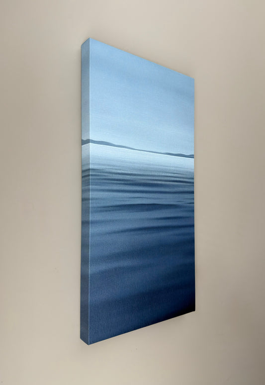 A vertical canvas wall art print depicting a calm blue ocean scene with horizon and soft wave textures, shown hanging on a beige wall.