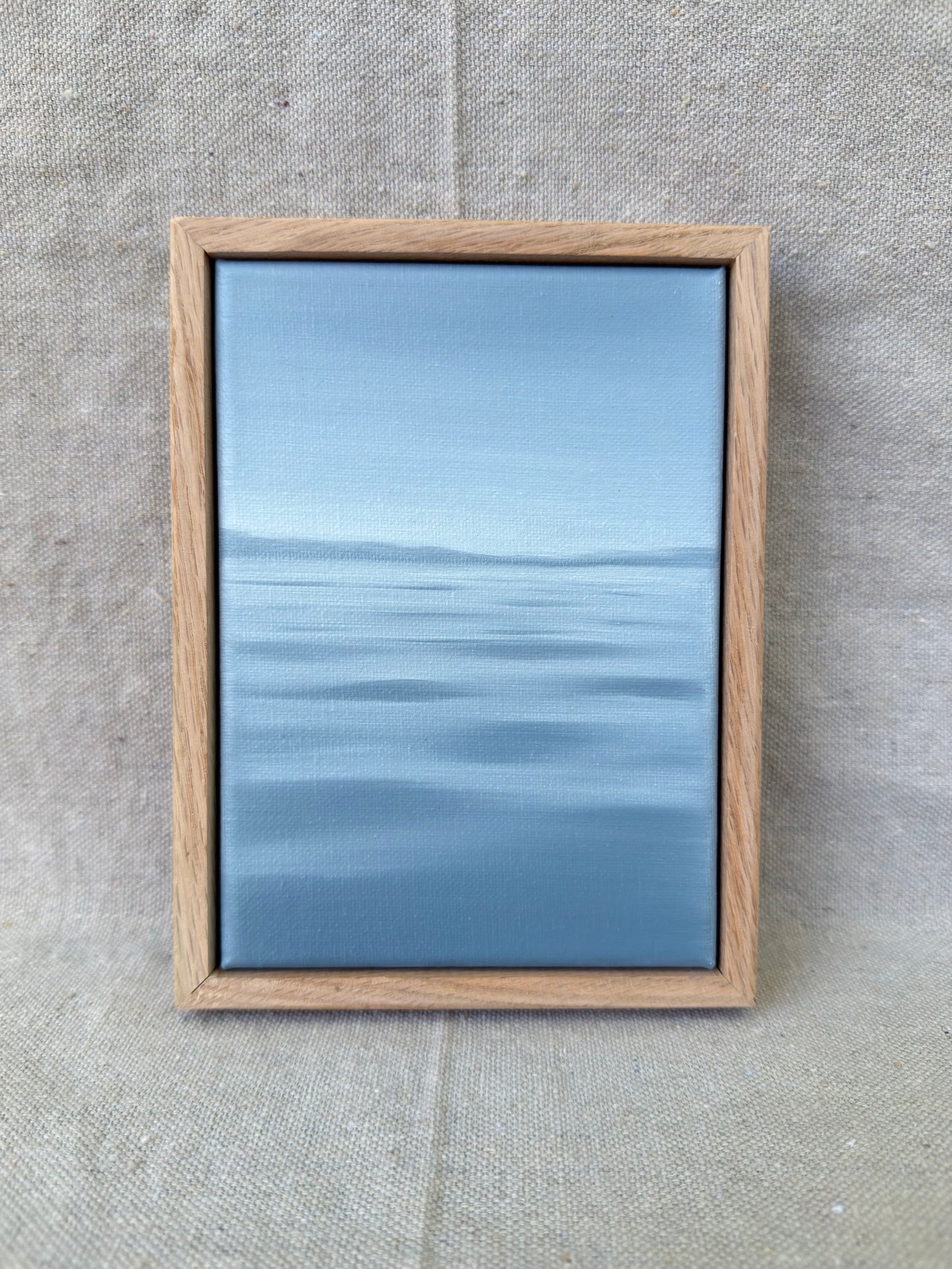 A framed minimalist landscape painting of a calm blue sea under a light sky, set in a light oak wood frame and displayed on a textured beige linen background.