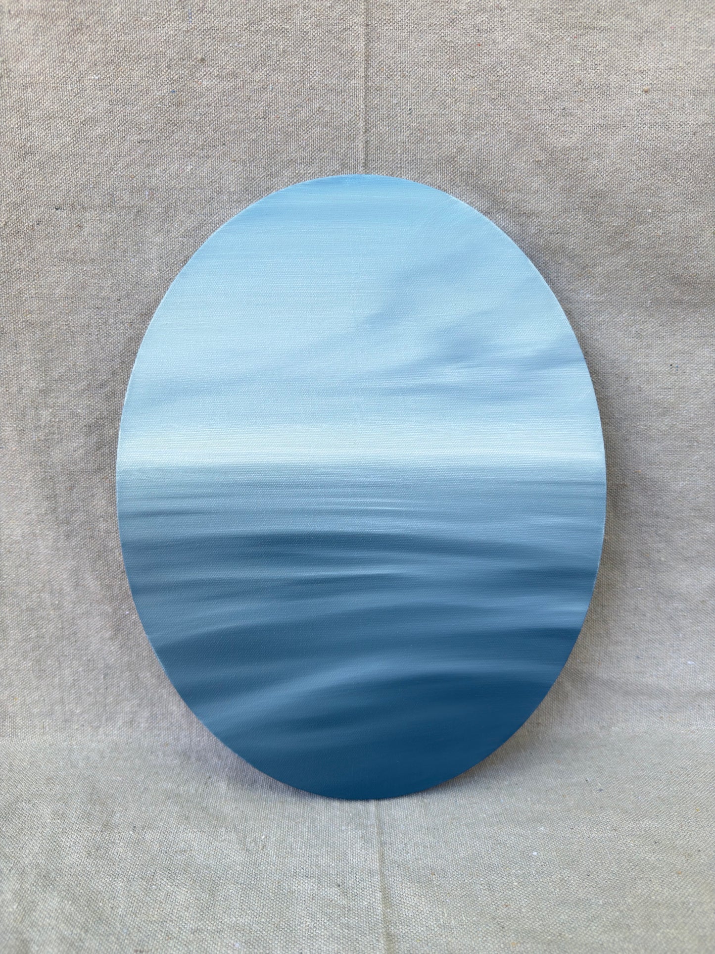 An oval canvas print depicting a serene blue ocean horizon under a light sky, displayed against a neutral beige textured fabric background.