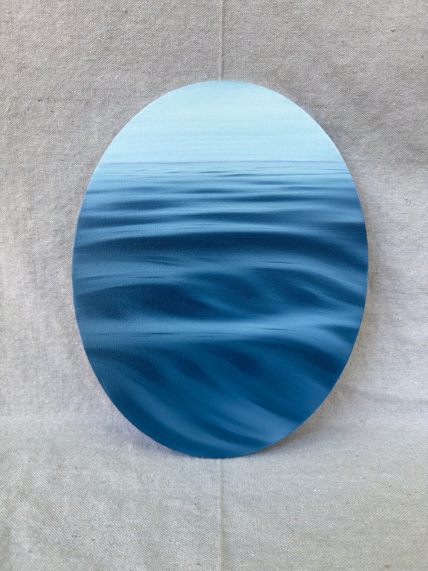 An oval wall art piece depicting a calm blue ocean with gentle waves under a pale sky, displayed against a neutral textured beige background.