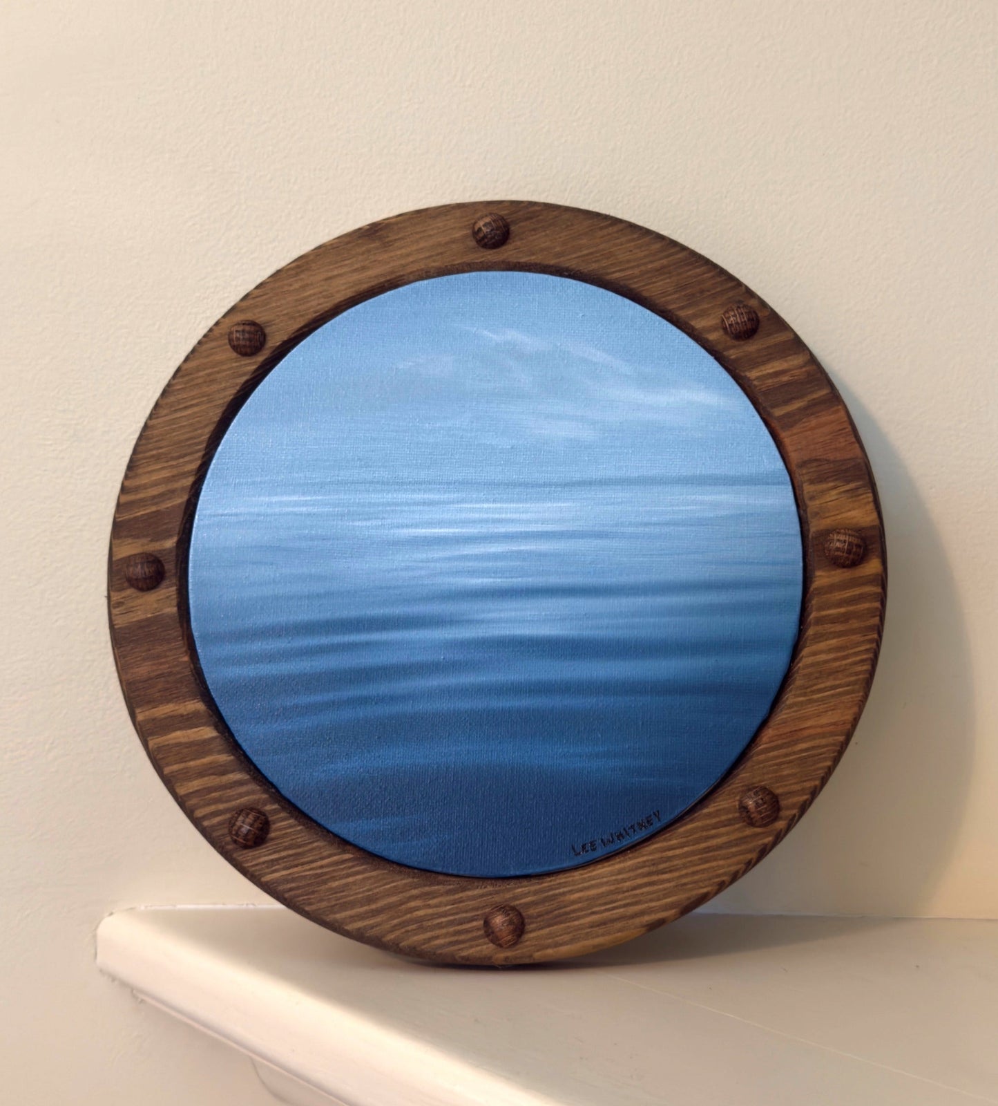Round ocean painting with wooden frame on a white shelf against a beige wall