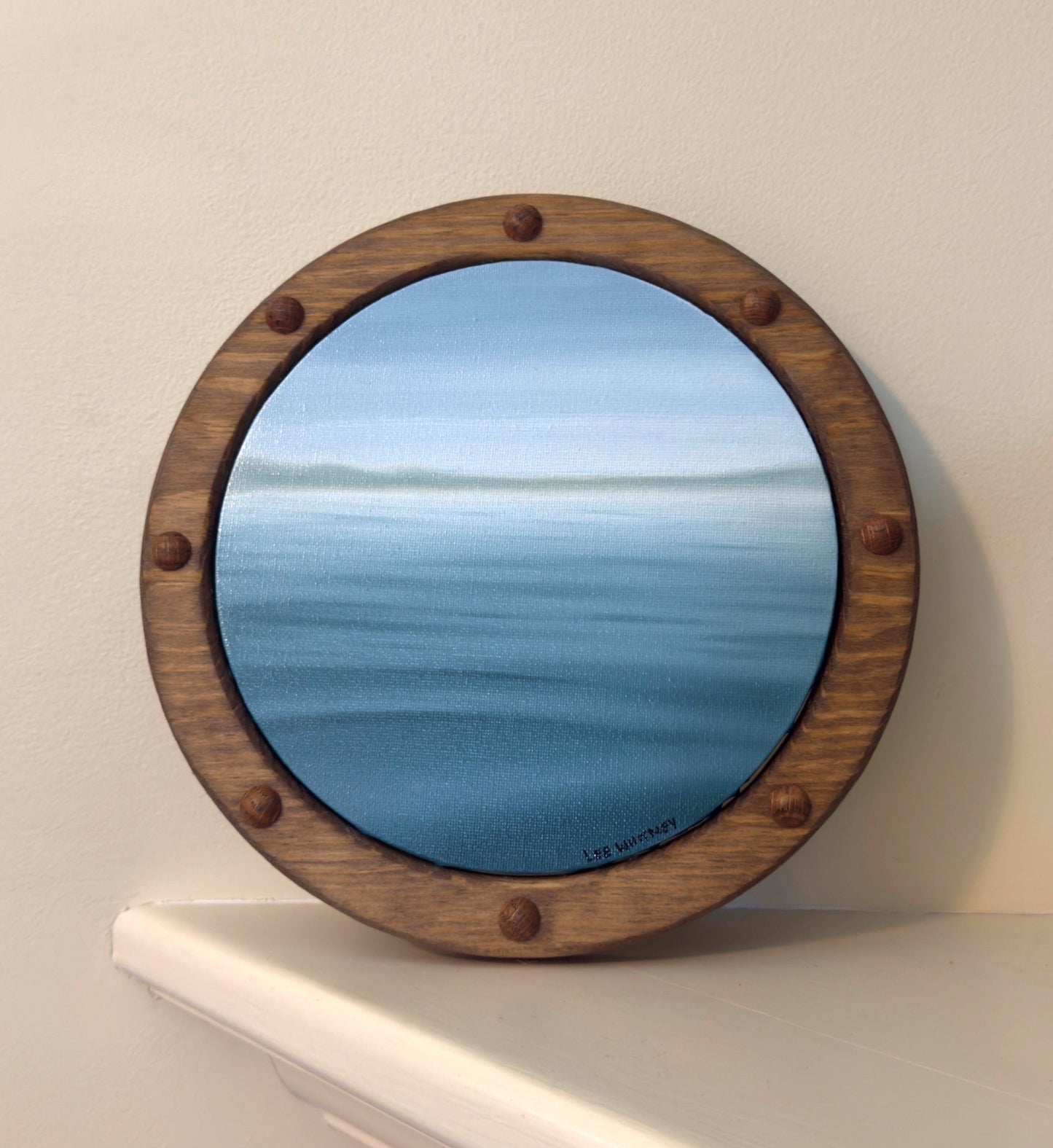 Round wooden-framed green water painting on a white shelf against a beige wall.