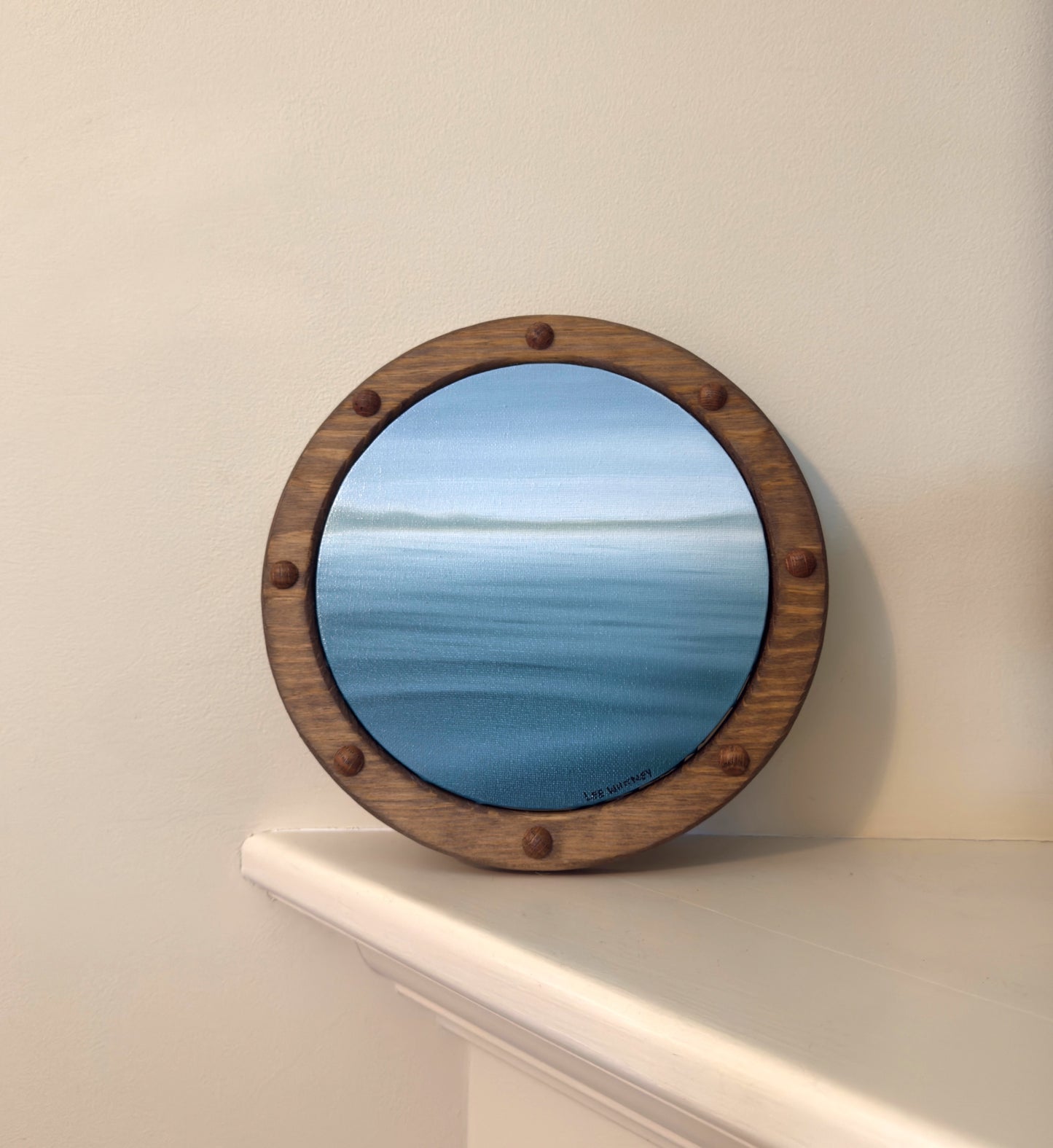 Round wooden-framed green water painting on a white shelf against a beige wall.