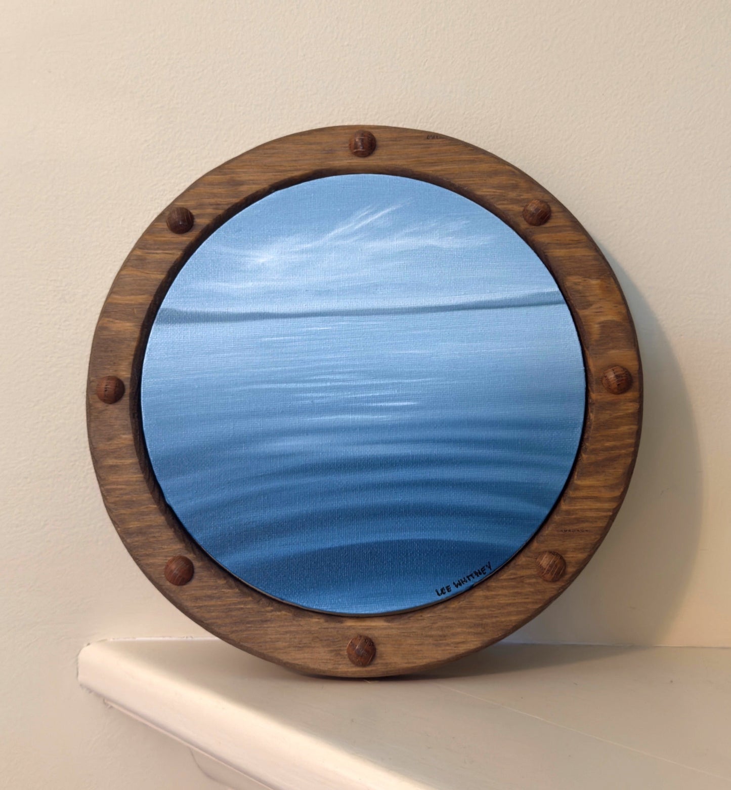 Round wooden-framed ocean painting on a white shelf against a beige wall