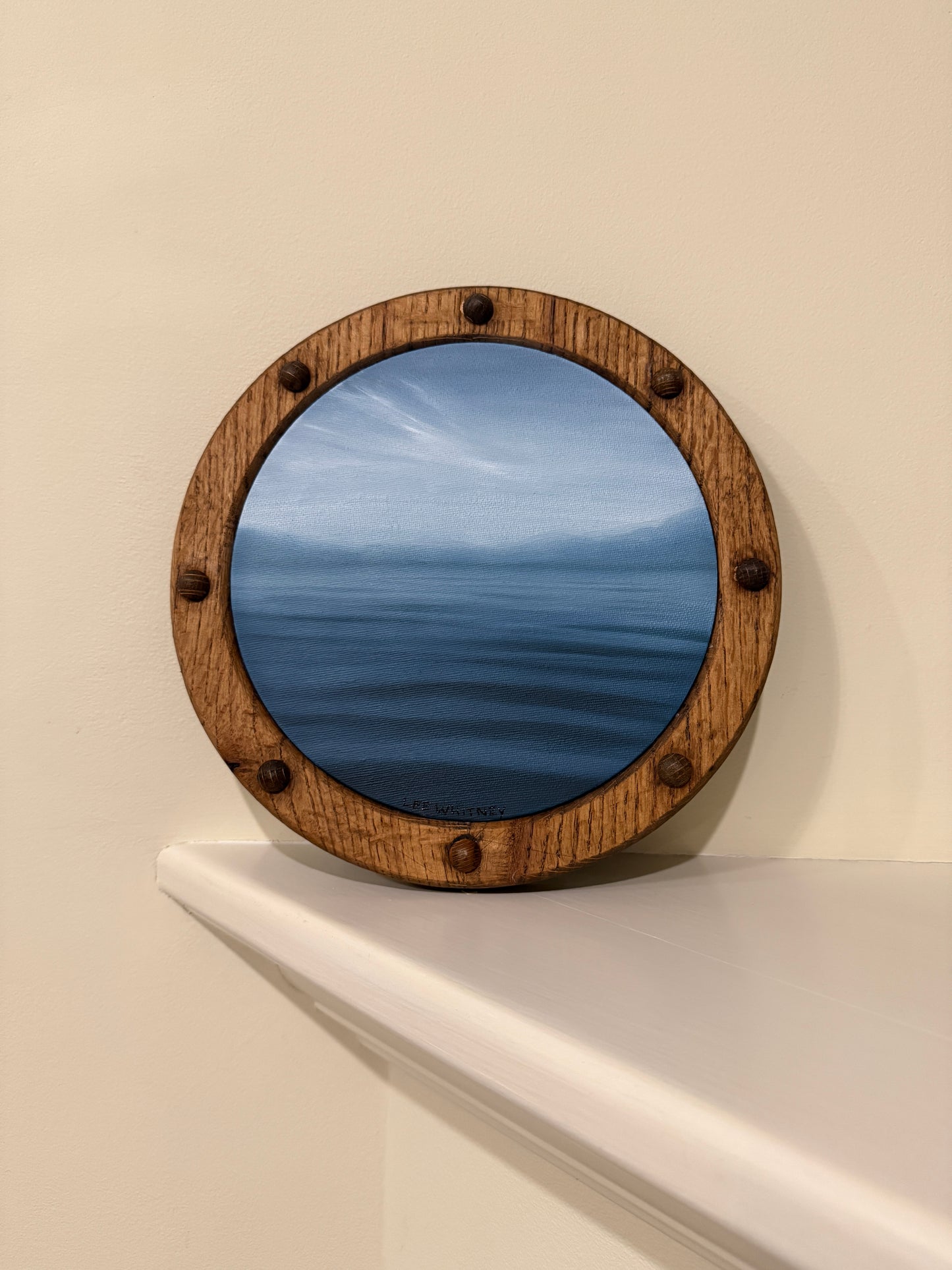 Round wooden-framed waterscape painting in slate blue on an off-white wall shelf