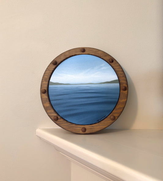 Round wooden-framed painting reflecting a scenic view on a beige wall.
