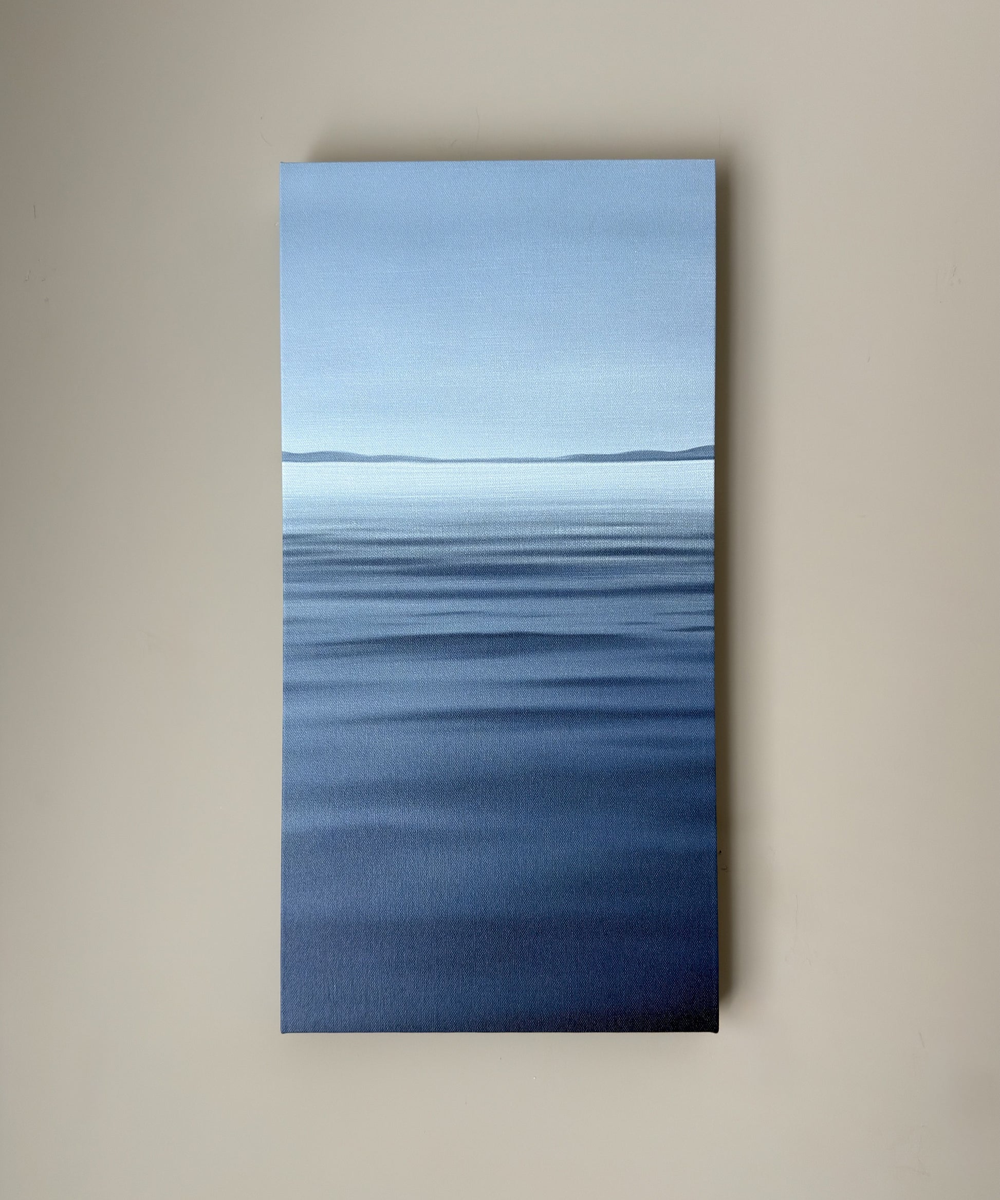 A vertical canvas wall art print depicting a calm blue ocean with a distant horizon under a pale sky, mounted on a beige wall.