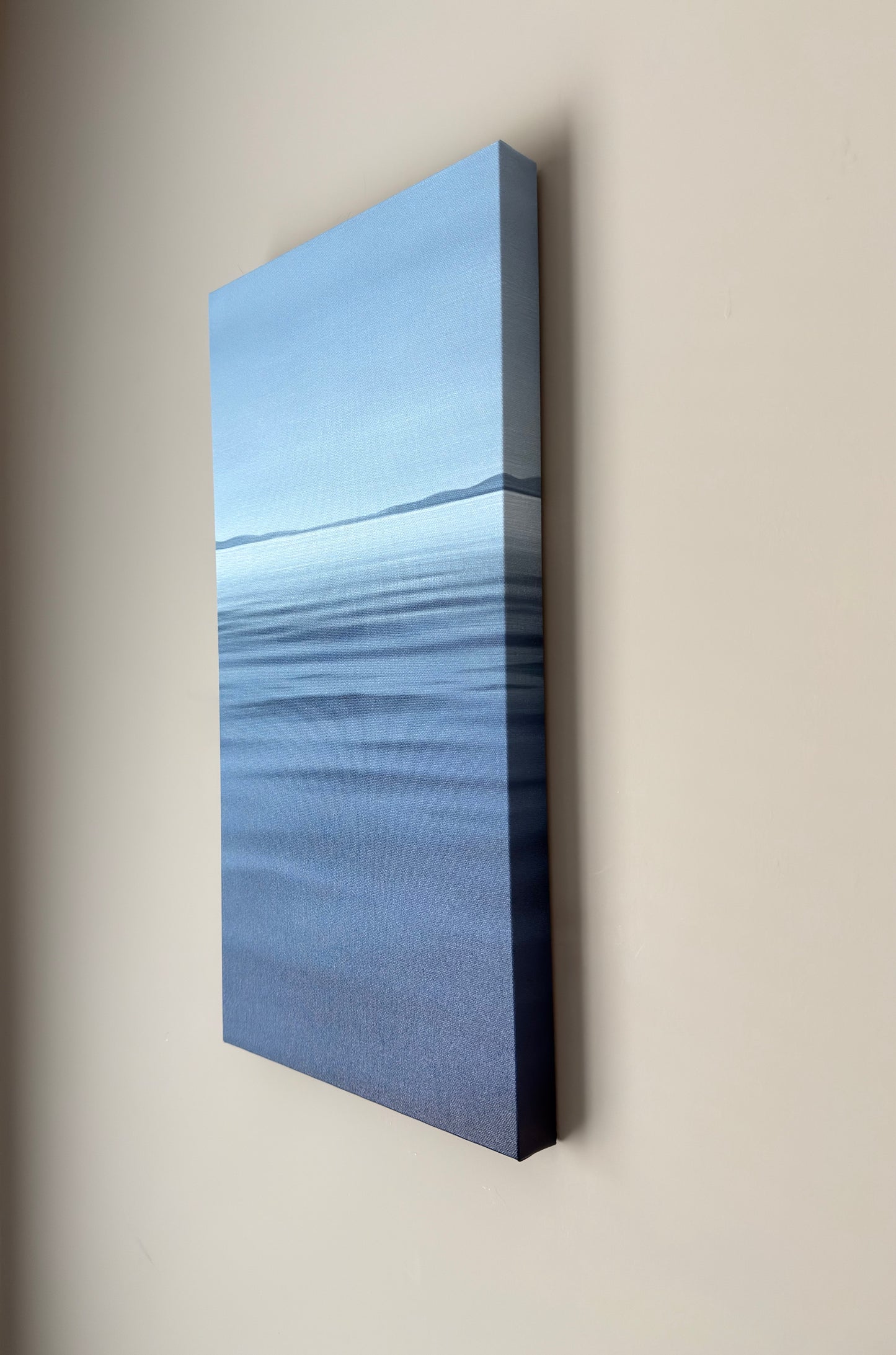 A vertical canvas wall art print depicting a calm blue water scene with a distant shoreline, mounted on a beige wall.
