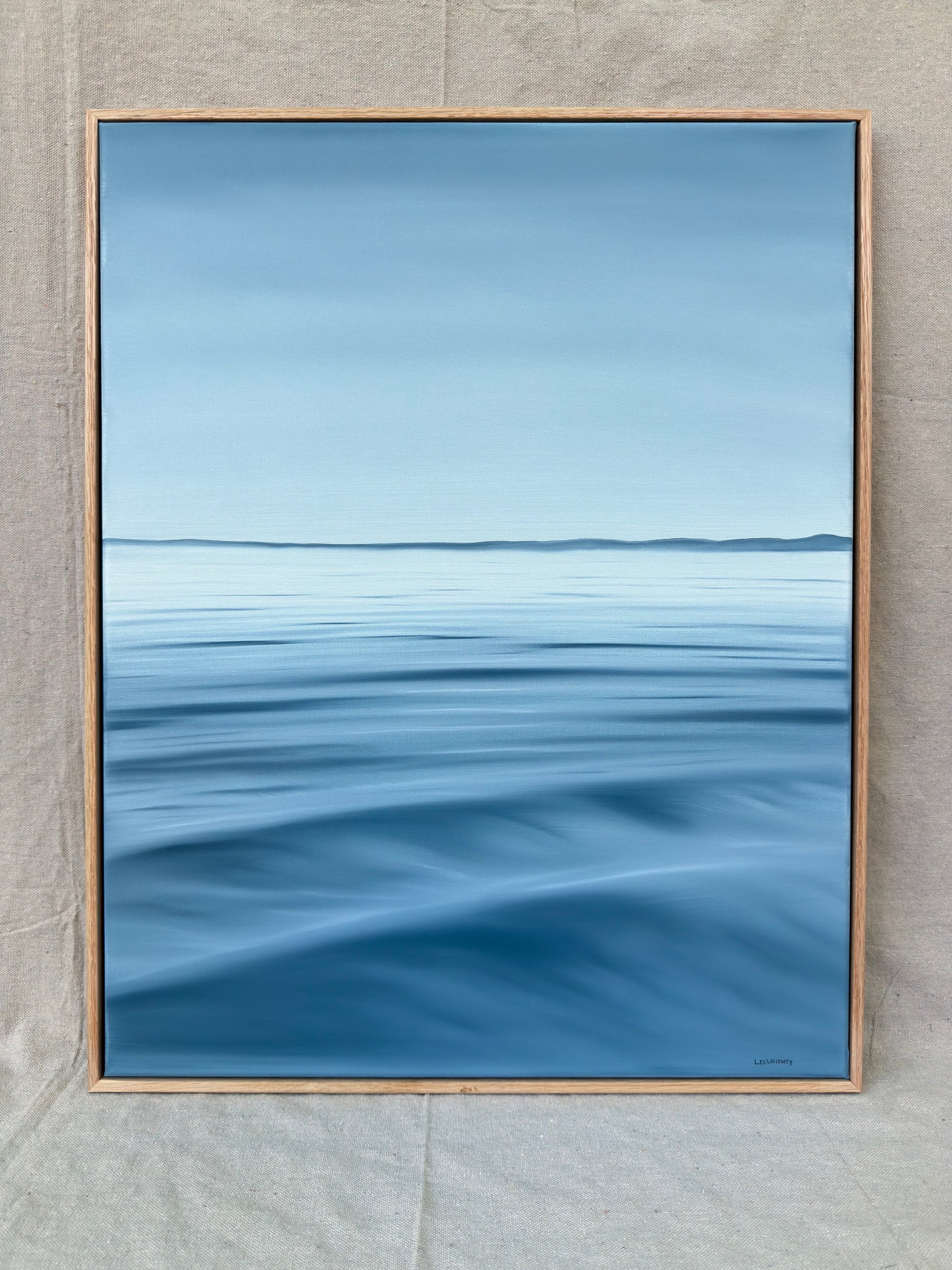 A framed landscape painting of a calm blue sea under a pale sky, displayed against a neutral textured gray fabric background.