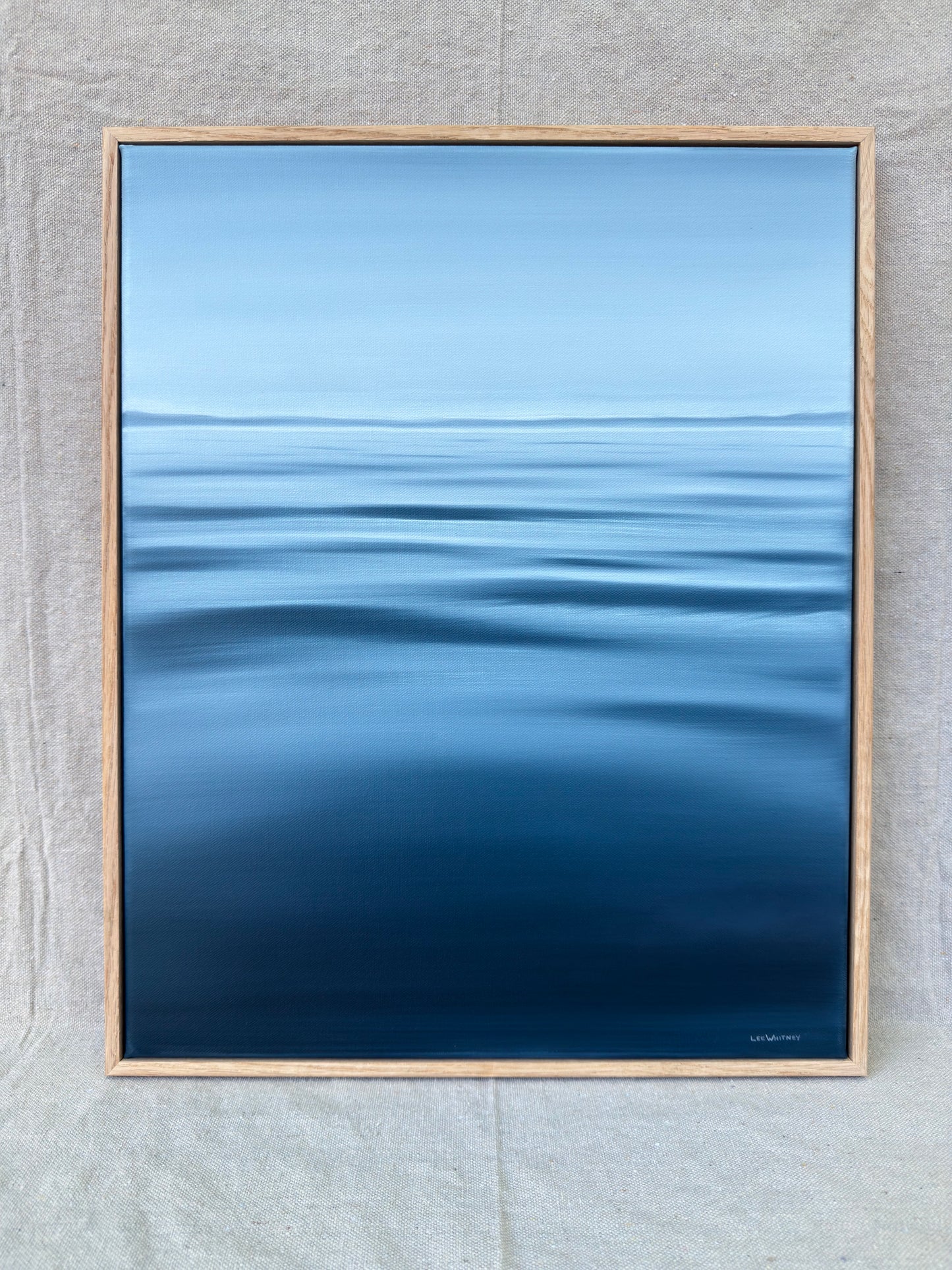 A framed art print depicting a serene blue ocean with gentle ripples under a light blue sky, set in a light wood frame against a textured gray fabric background.