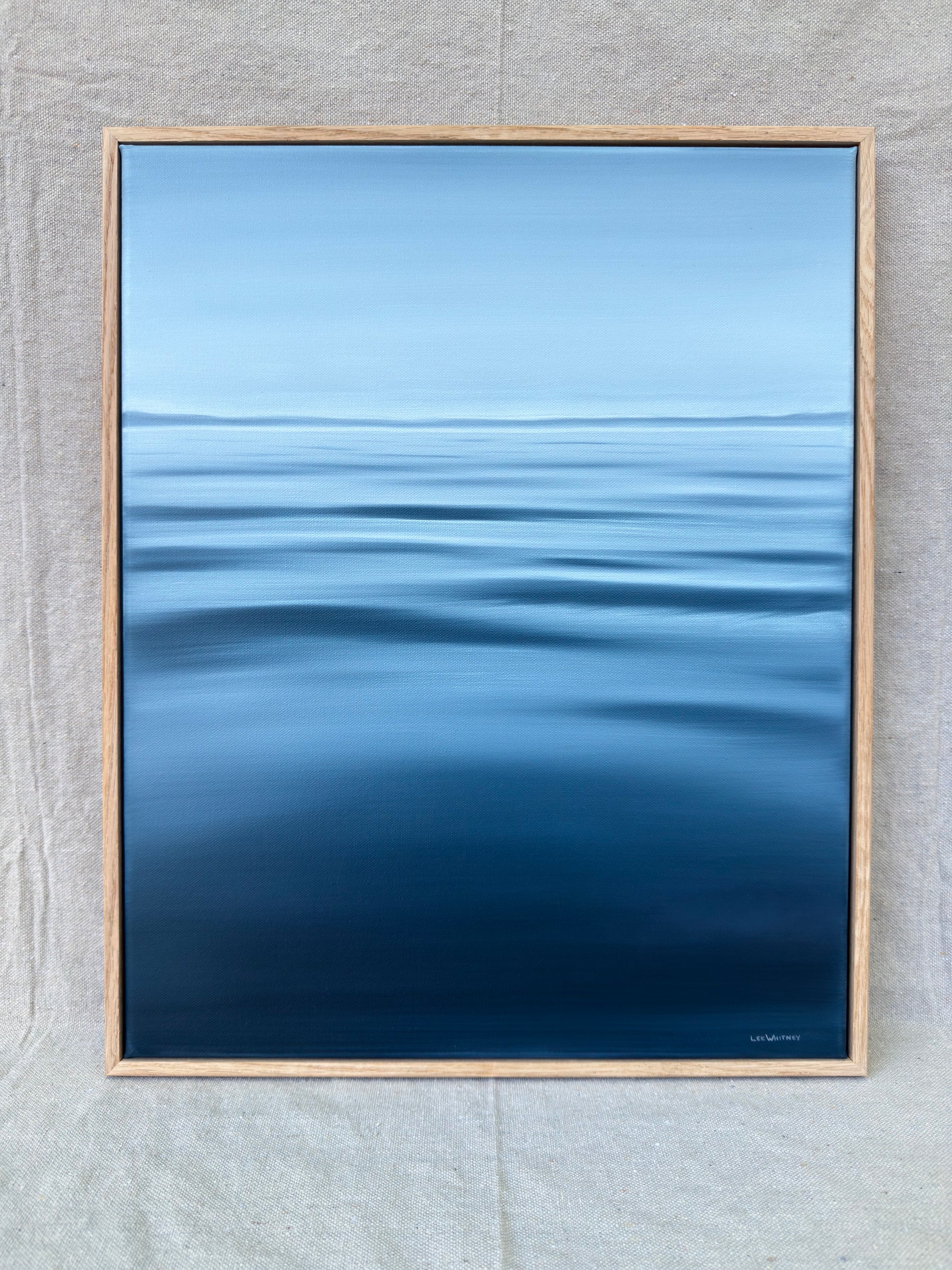 A framed art print depicting a serene blue ocean with gentle ripples under a light blue sky, set in a light wood frame against a textured gray fabric background.