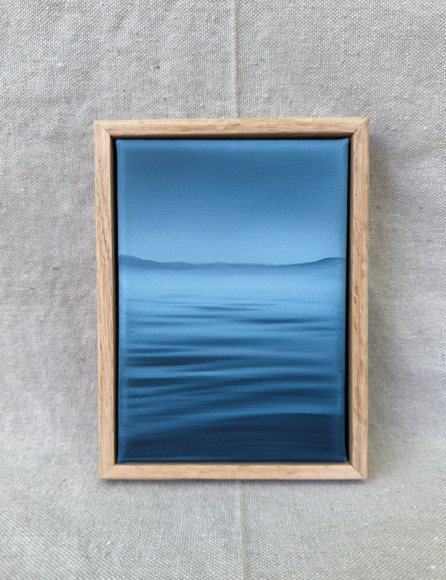 A framed art print featuring a serene blue landscape with calm water and distant hills, enclosed in a light oak wood frame and displayed against a textured beige linen background.