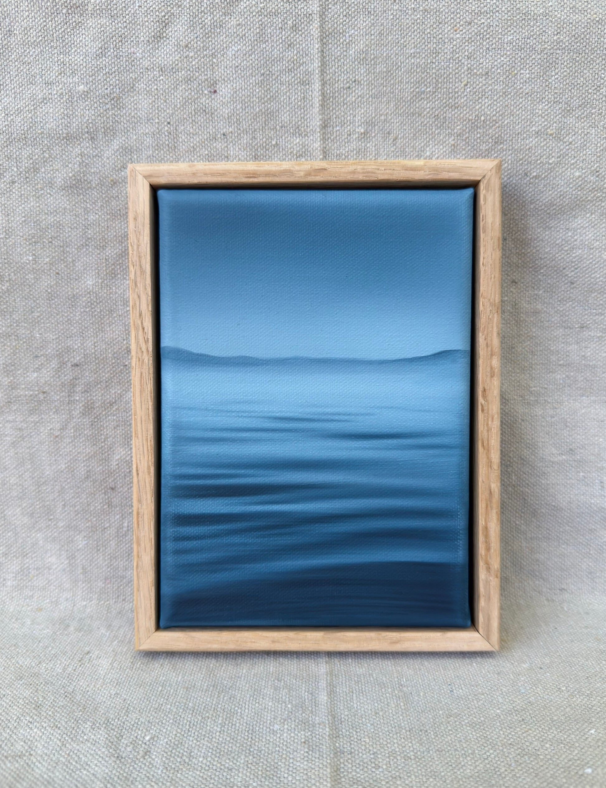 A framed art print featuring a serene blue landscape with calm water and distant hills, enclosed in a light oak wood frame and displayed against a textured beige linen background.