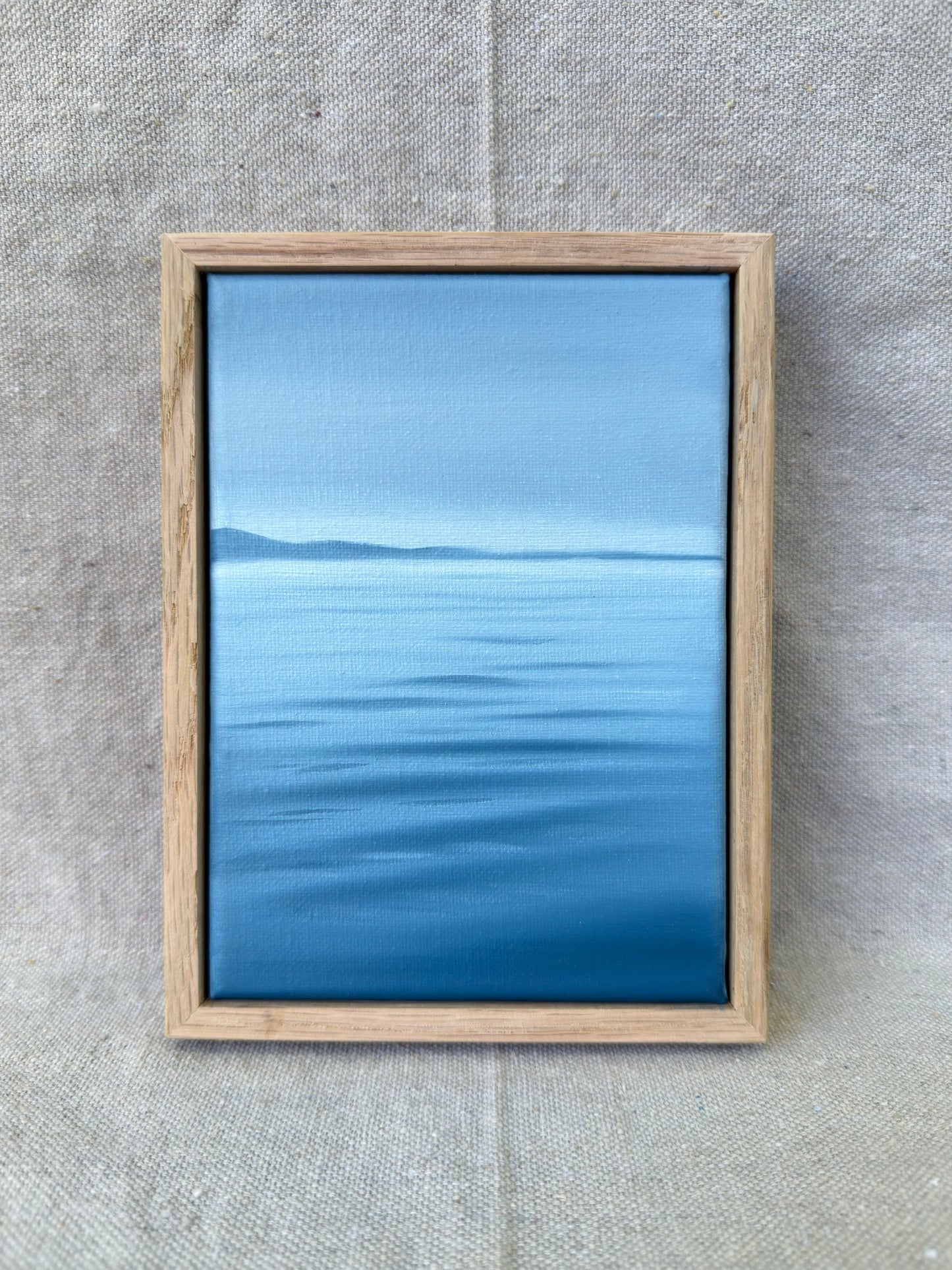A framed landscape painting of a calm blue sea under a clear sky, set in a light natural wood frame and displayed on a textured beige linen background.