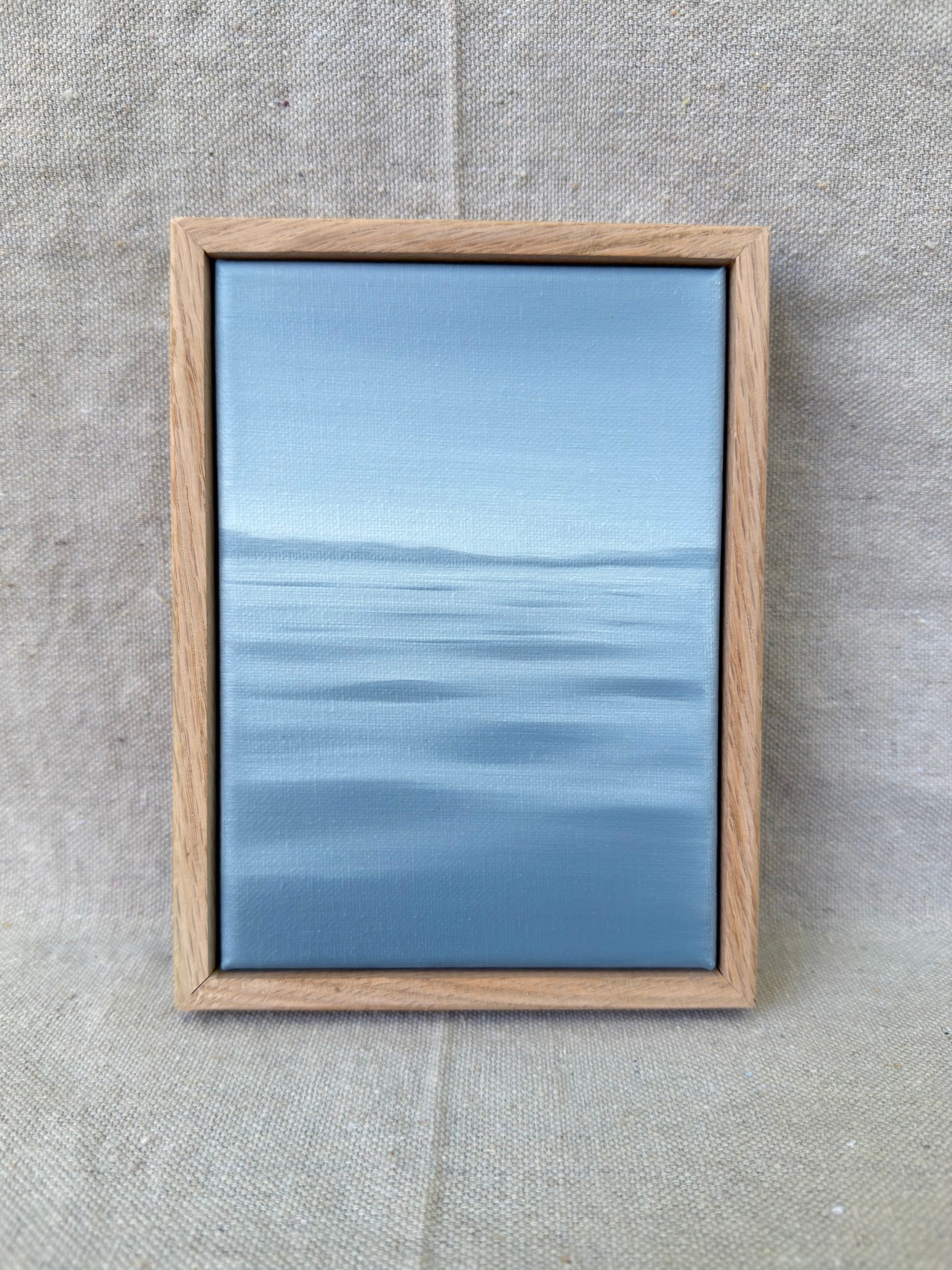 A framed minimalist landscape painting of a calm blue sea under a light sky, set in a light oak wood frame and displayed on a textured beige linen background.