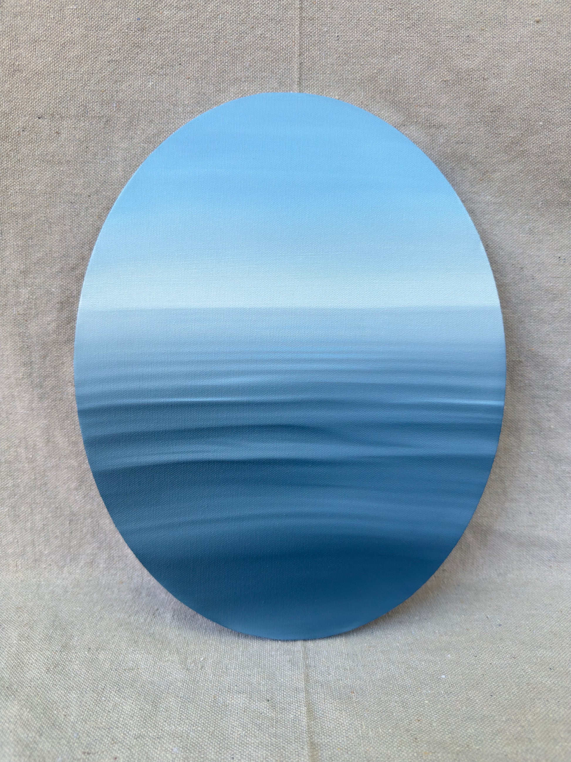 An oval canvas print depicting a calm blue ocean horizon, displayed against a neutral beige textured fabric background.