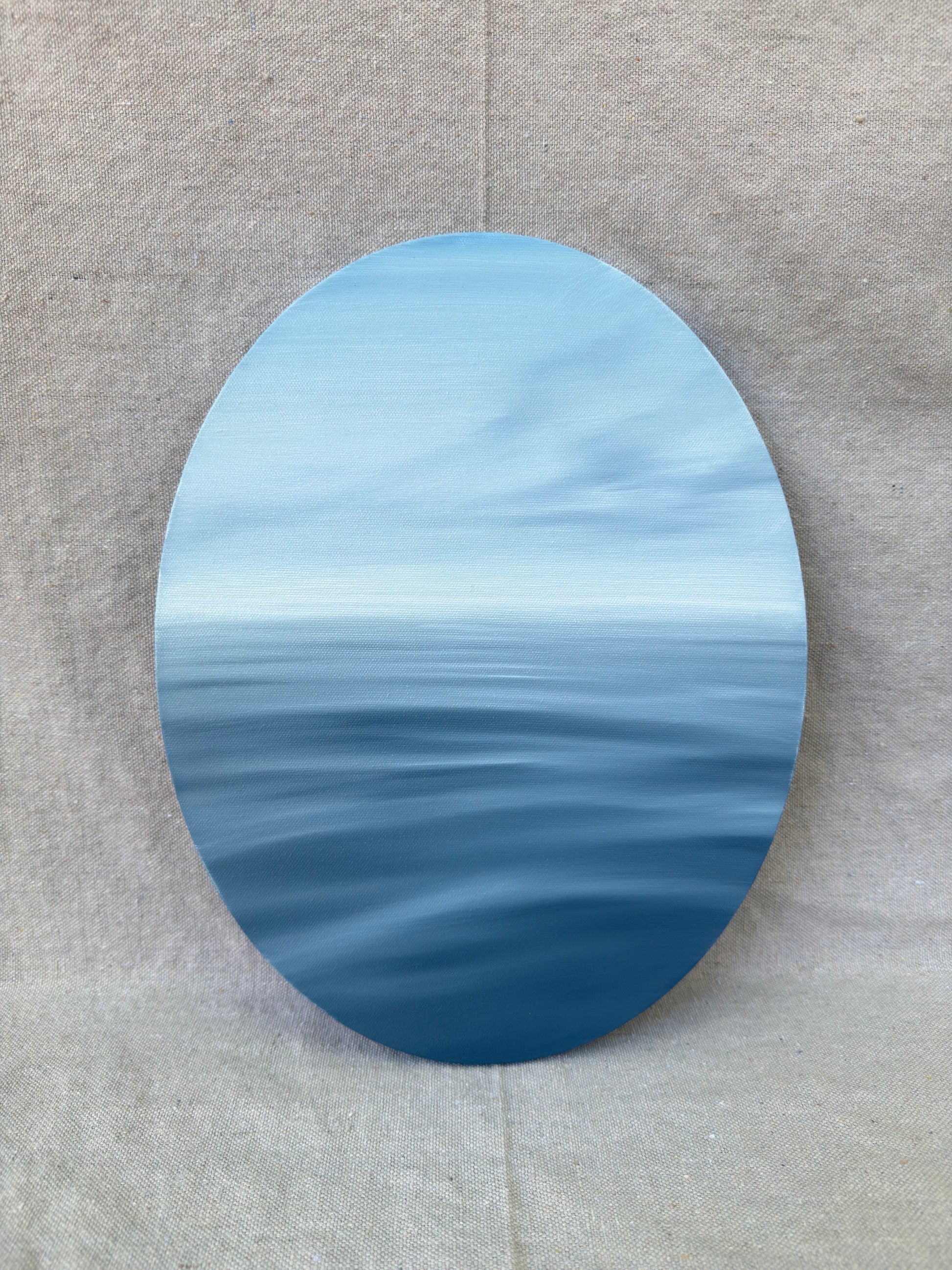 An oval canvas print depicting a serene blue ocean horizon under a light sky, displayed against a neutral beige textured fabric background.