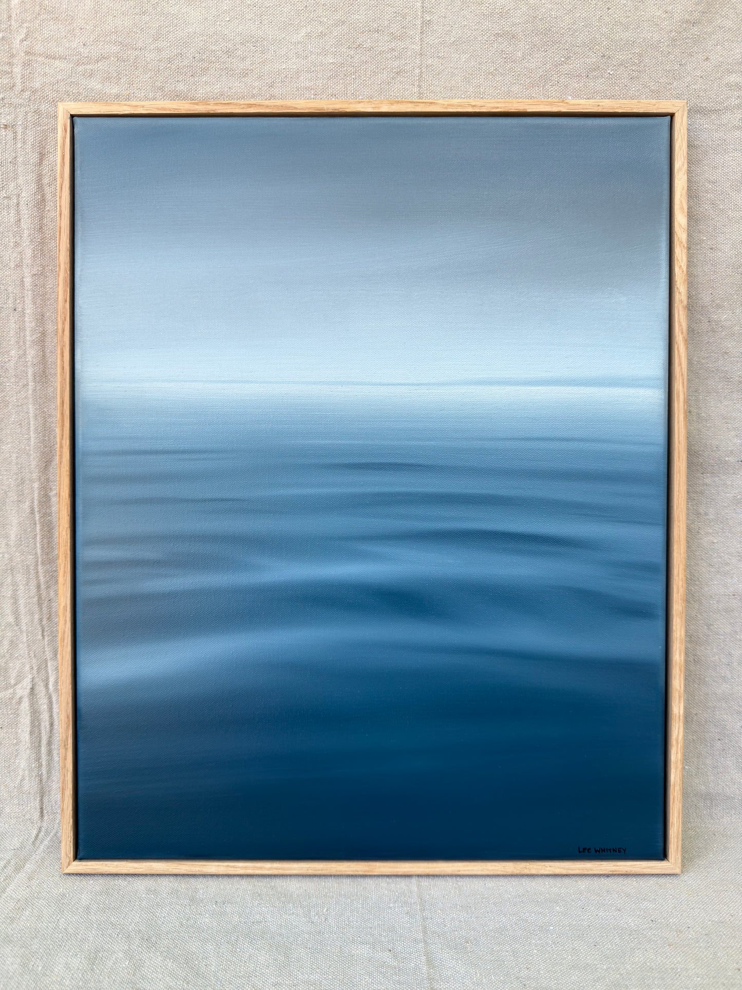 A framed abstract seascape painting featuring a gradient of blue and gray tones representing water and sky, enclosed in a light natural wood frame and displayed against a textured beige wall.