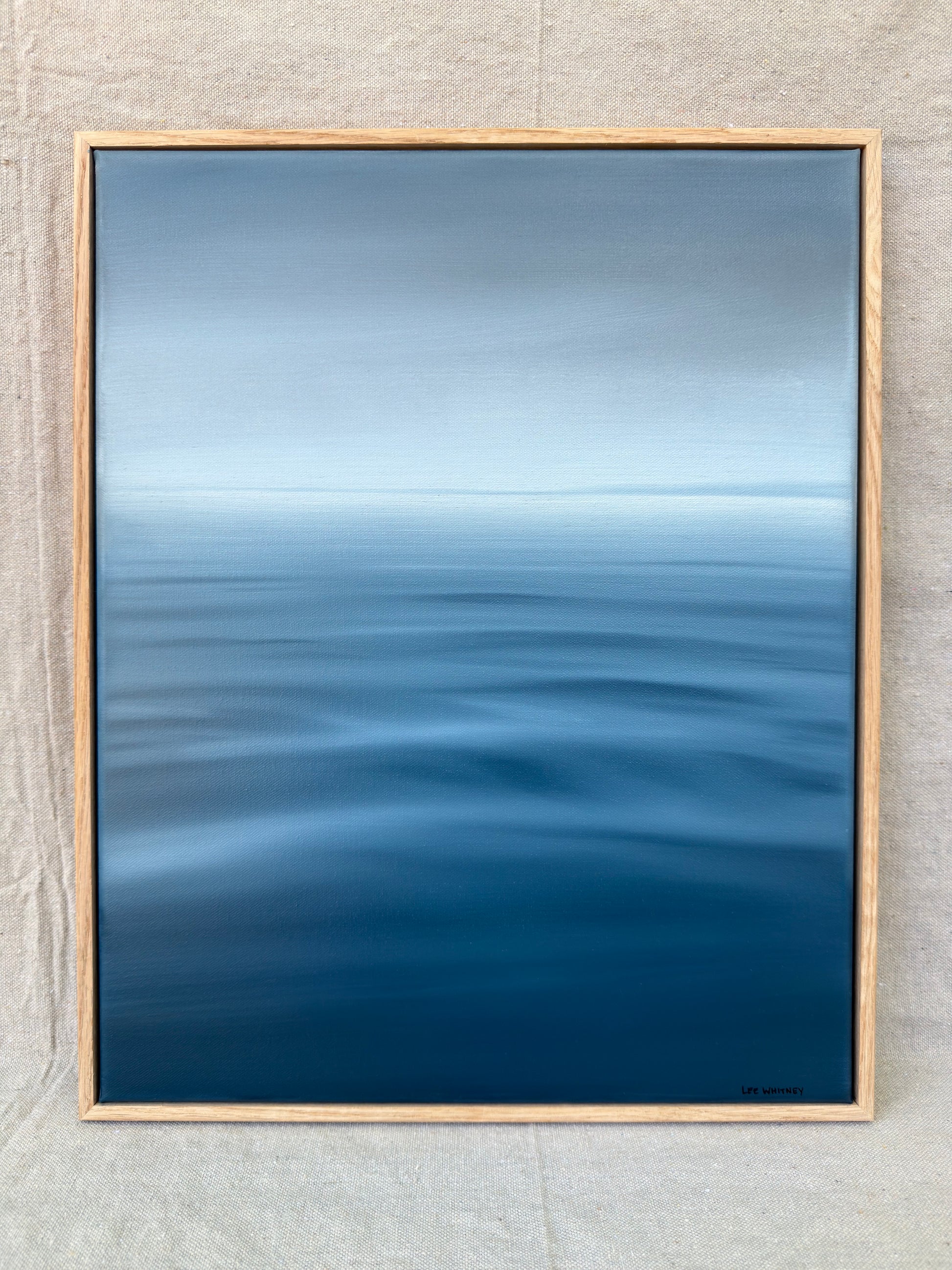 A framed abstract seascape painting featuring a gradient of blue and gray tones representing water and sky, enclosed in a light natural wood frame and displayed against a textured beige wall.