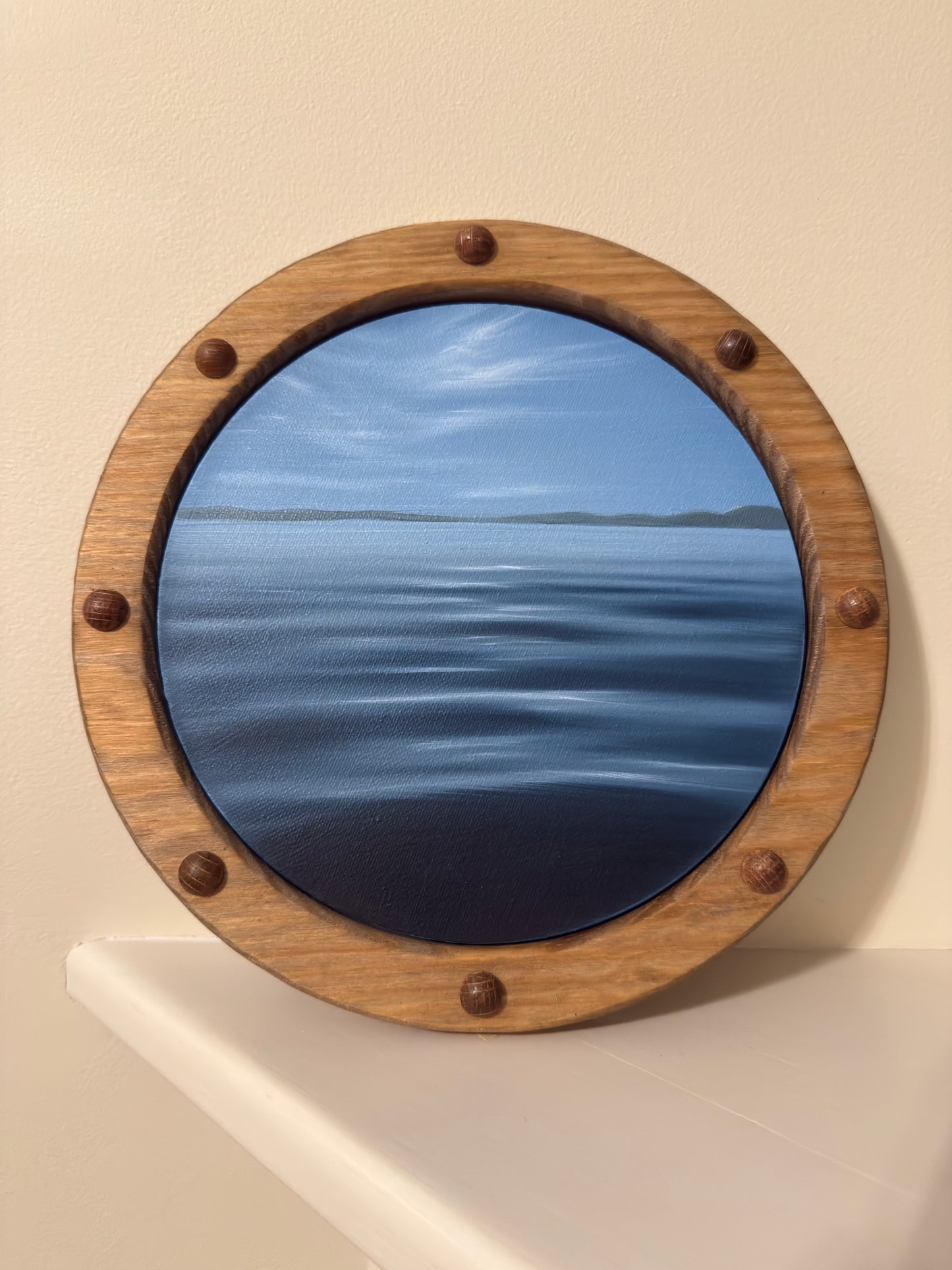 A circular porthole-style framed artwork showing a blue ocean and sky scene, mounted on a beige wall above a white ledge. The frame is made of medium brown wood with six dark metal rivets.
