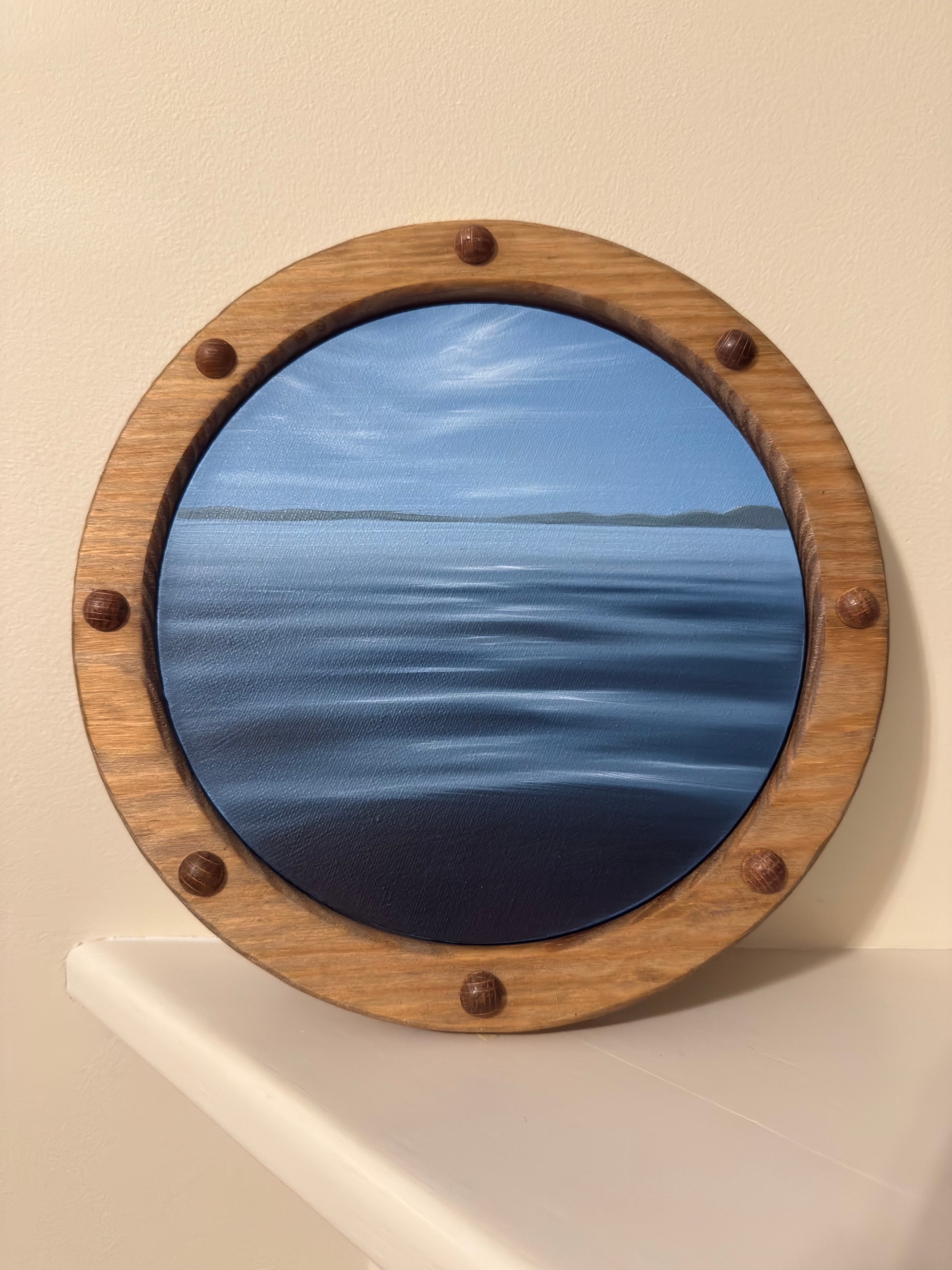 A circular porthole-style framed artwork showing a blue ocean and sky scene, mounted on a beige wall above a white ledge. The frame is made of medium brown wood with six dark metal rivets.