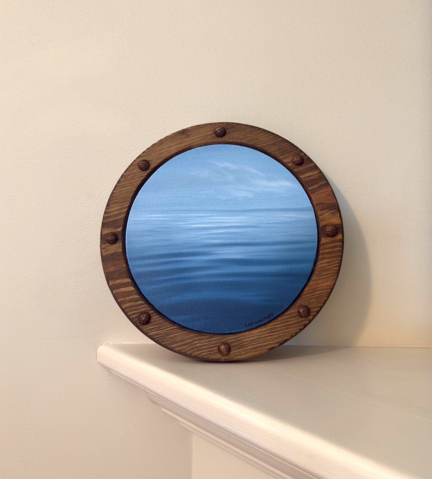 Round ocean painting with wooden frame on a white shelf against a beige wall