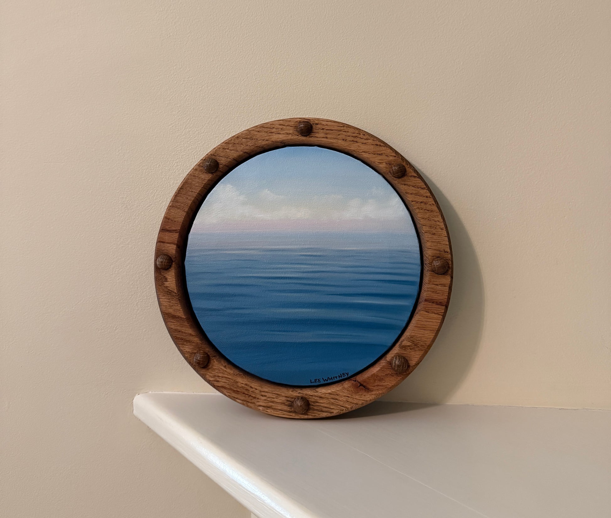 Round wooden-framed waterscape painting reflecting a blue ocean scene on an off-white wall shelf