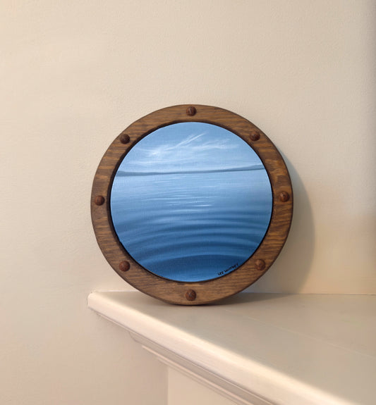 Round wooden-framed ocean painting on a white shelf against a beige wall