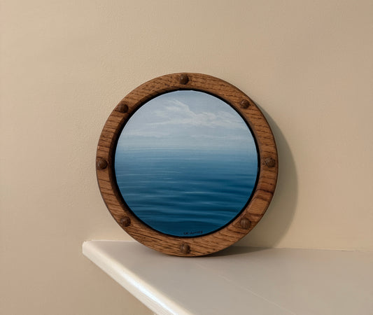 Round wooden-framed daytime waterscape painting on an off-white wall shelf