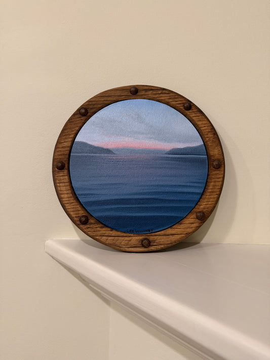 Round wooden framed pink sunset waterscape painting on an off-white wall shelf