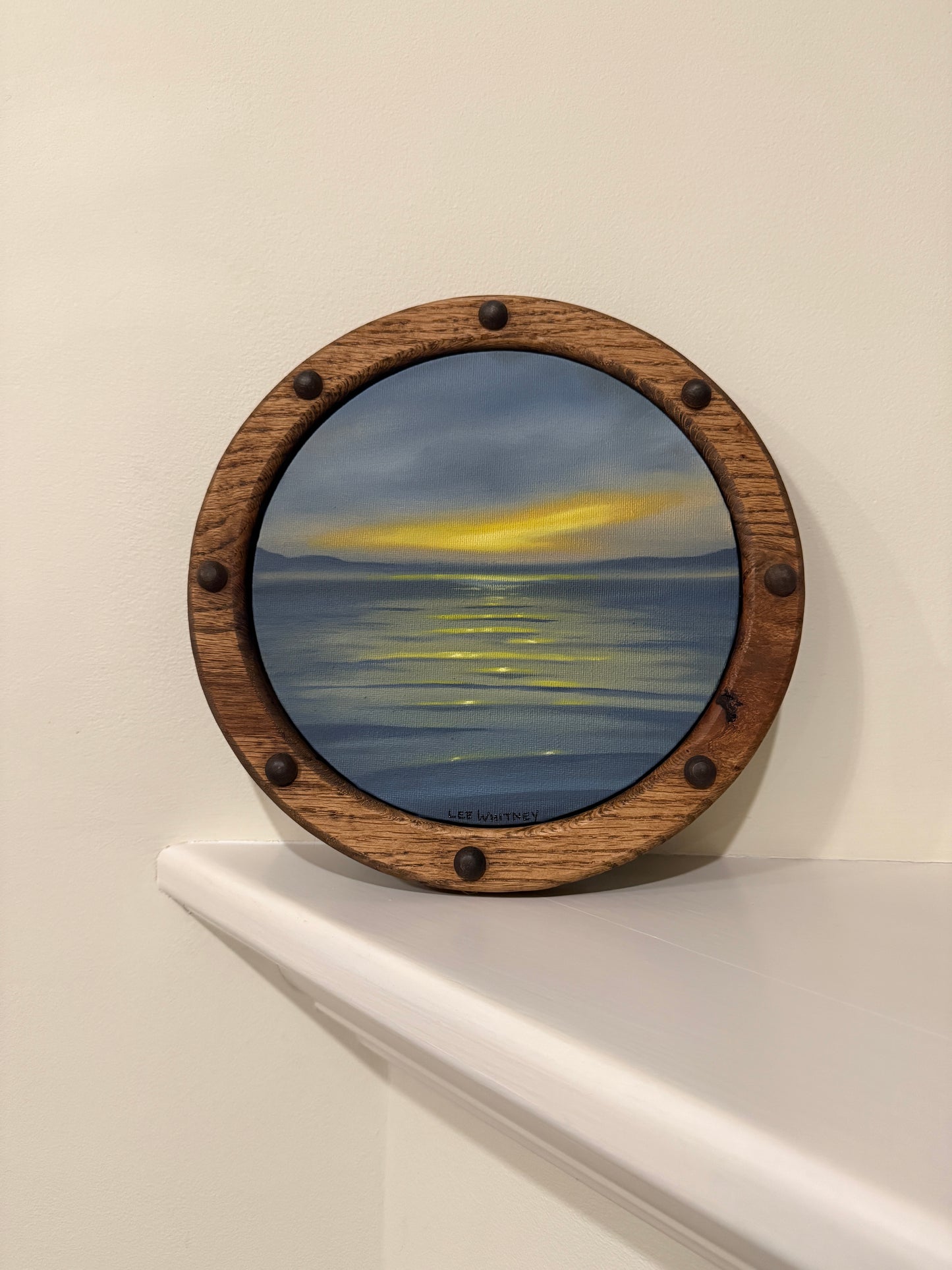 Round wooden frame with a sunset waterscape painting on an off-white wall shelf
